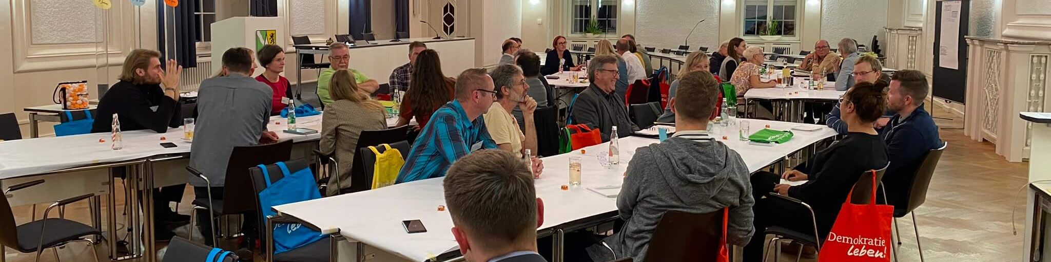 KI generiert: Das Bild zeigt eine Gruppe von Menschen, die in einem hellen Raum mit langen Tischen sitzen und sich unterhalten. Der Hauptinhalt des Bildes ist eine Versammlung oder ein Treffen, möglicherweise eine Konferenz oder ein Workshop.