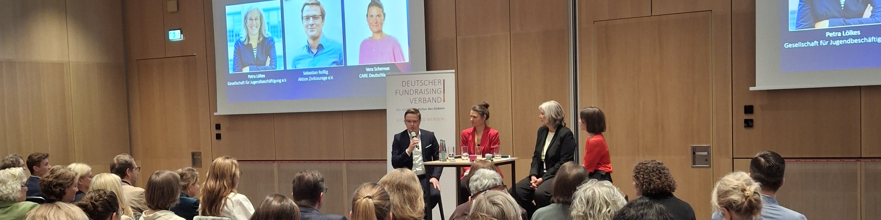 KI generiert: Das Bild zeigt eine Podiumsdiskussion mit mehreren Teilnehmern auf einer Bühne, die vor einem Publikum stattfindet. Im Hintergrund sind eine Leinwand mit den Namen und Titeln der Diskutanten sowie das Logo des Deutschen Fundraising Verbands zu sehen.