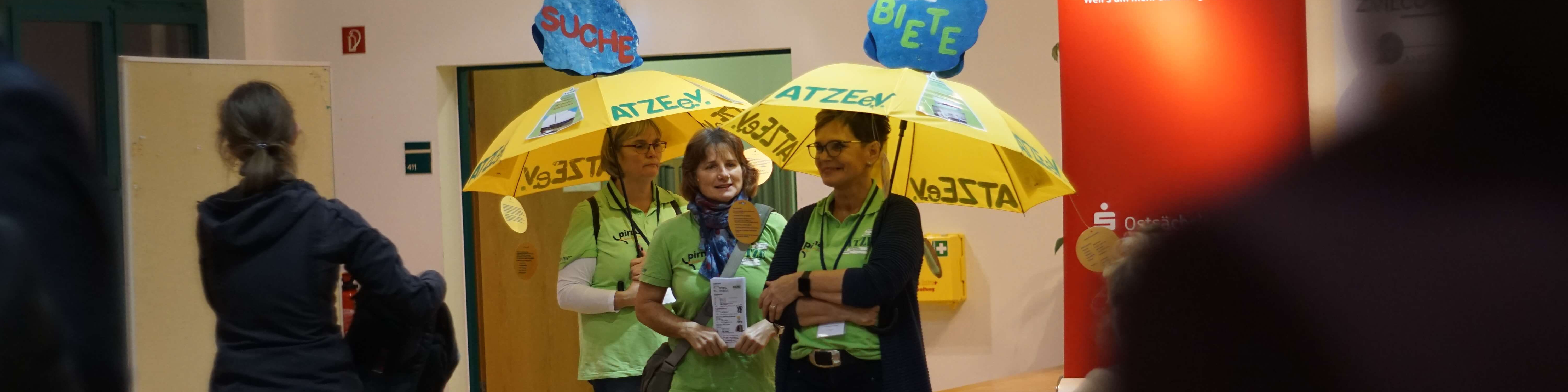 KI generiert: Das Bild zeigt drei Personen, die gelbe Regenschirme mit der Aufschrift "ATZEN" und "No3STA" halten, in einem Innenraum, möglicherweise bei einer Veranstaltung oder Demonstration. Im Hintergrund sind weitere Personen und ein rotes Banner zu sehen.
