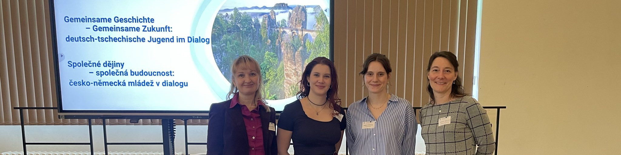 KI generiert: Das Bild zeigt vier Personen, die vor einer Leinwand mit einer Präsentation stehen. Die Präsentation hat den Titel "Gemeinsame Geschichte - Gemeinsame Zukunft: deutsch-tschechische Jugend im Dialog" und die tschechische Übersetzung darunter.