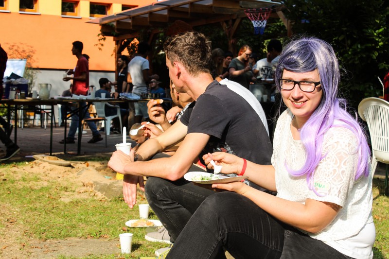 KI generiert: Das Bild zeigt eine Gruppe von Menschen, die draußen an Tischen sitzen und gemeinsam essen, während im Hintergrund weitere Personen stehen und sich unterhalten. Der Hauptinhalt des Bildes ist eine gesellige Zusammenkunft oder ein Picknick in einer entspannten Atmosphäre.