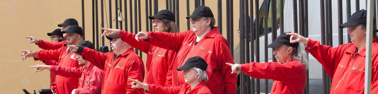 KI generiert: Das Bild zeigt eine Gruppe von Personen, die in roten Uniformen und schwarzen Mützen gekleidet sind und synchronisierte Armbewegungen ausführen. Der Hauptinhalt des Bildes ist eine choreografierte Darbietung oder Übung im Freien.