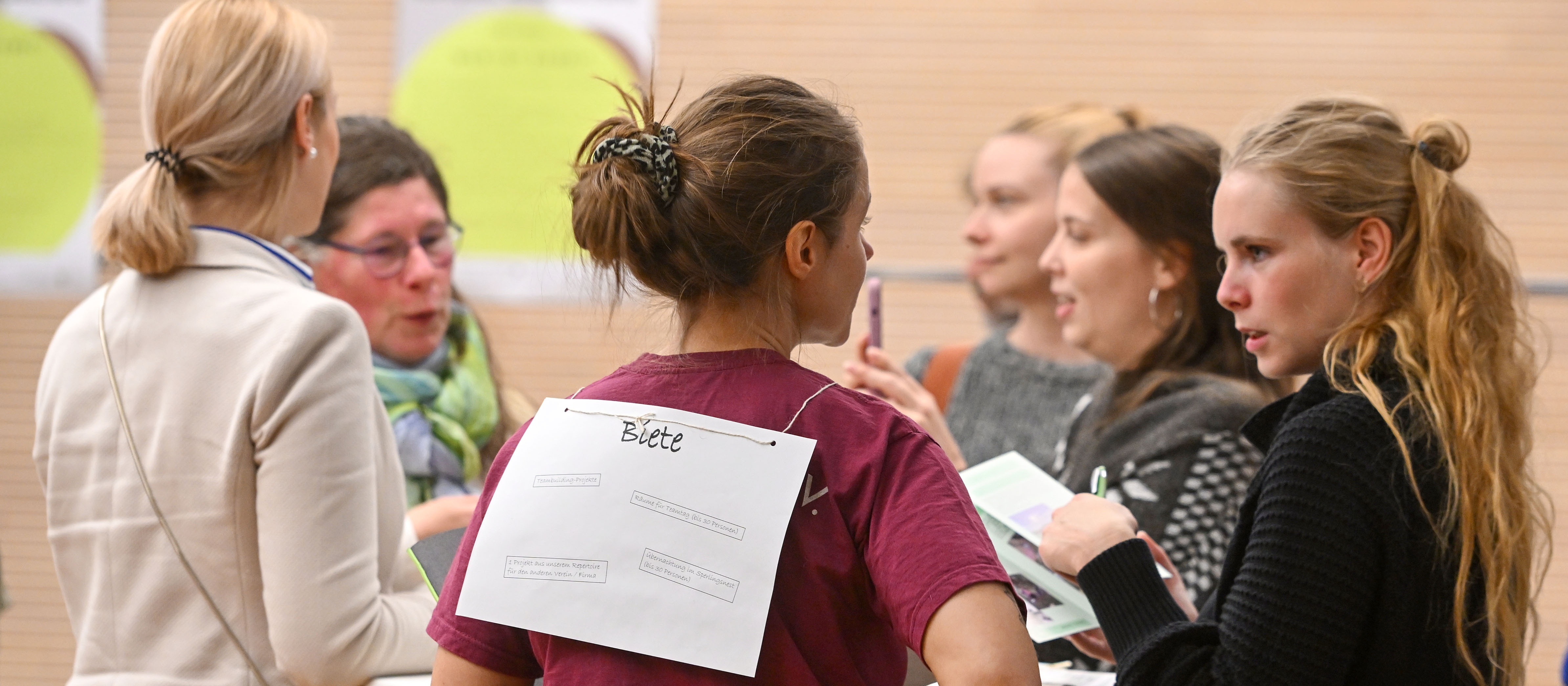 KI generiert: Das Bild zeigt eine Gruppe von Personen, die in einer informellen Besprechung oder einem Workshop zusammenstehen und sich unterhalten. Eine Person im Vordergrund hat ein Blatt Papier mit der Aufschrift "Blatt" auf dem Rücken befestigt.