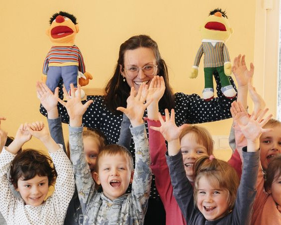 KI generiert: Das Bild zeigt eine Gruppe fröhlicher Kinder, die zusammen mit einer Erwachsenen in einem hellen Raum stehen und ihre Hände in die Luft strecken. Im Hintergrund sind zwei Puppen an der Fensterbank platziert.