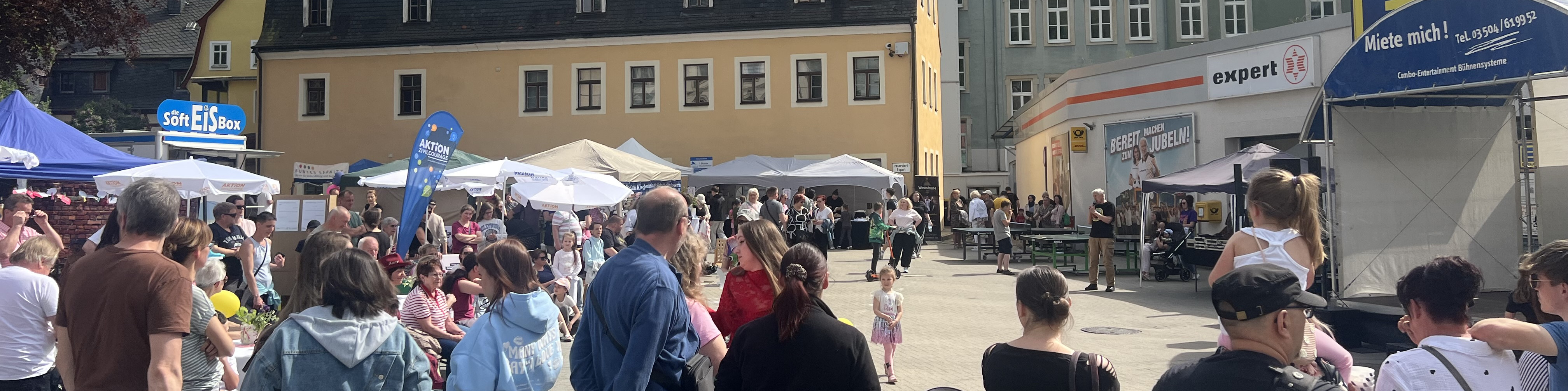 KI generiert: Das Bild zeigt eine belebte Straßenveranstaltung mit vielen Menschen, die an Ständen und unter Pavillons zusammenkommen. Im Hintergrund sind Gebäude und ein Werbeplakat zu sehen.