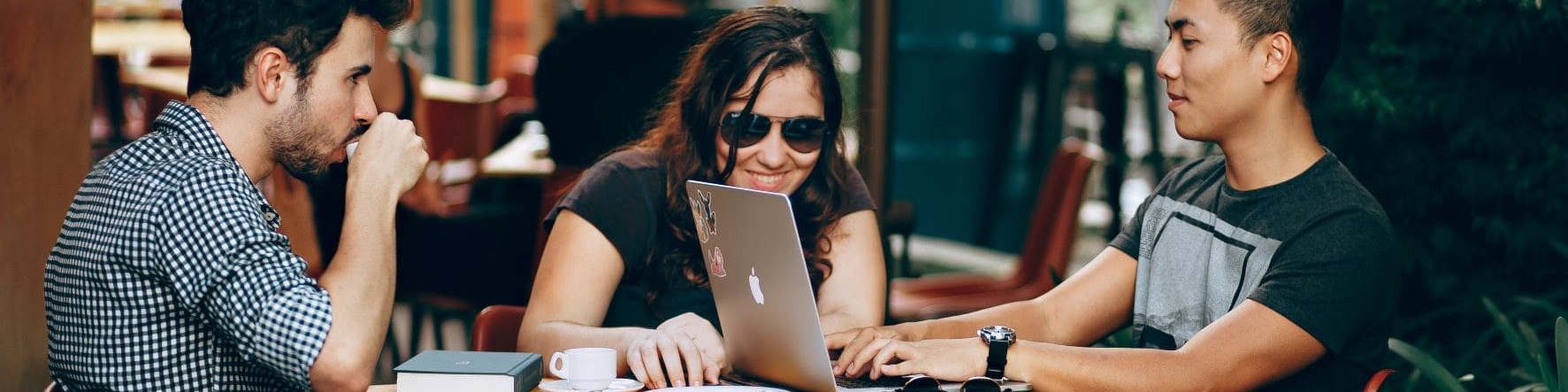 KI generiert: Das Bild zeigt drei Personen, die an einem Tisch sitzen und gemeinsam an einem Laptop arbeiten, vermutlich in einem Café oder einer informellen Arbeitsumgebung. Der Hauptinhalt des Bildes ist die Zusammenarbeit und Diskussion der Gruppe.