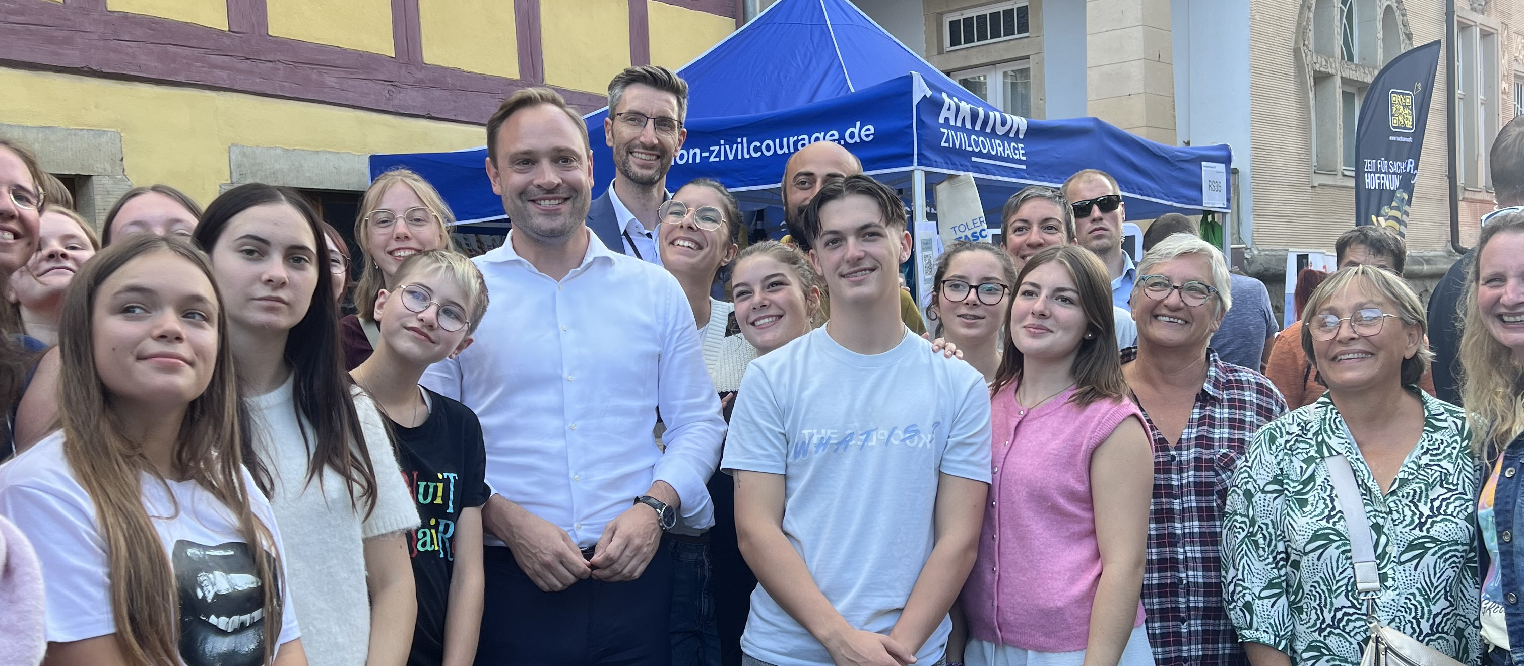 KI generiert: Das Bild zeigt eine Gruppe von Menschen, die sich vor einem blauen Zelt mit der Aufschrift "Aktion Zivilcourage" versammelt haben. Der Hauptinhalt des Bildes ist eine öffentliche Veranstaltung oder ein Treffen, das sich auf zivilen Mut und gesellschaftliches Engagement konzentriert.
