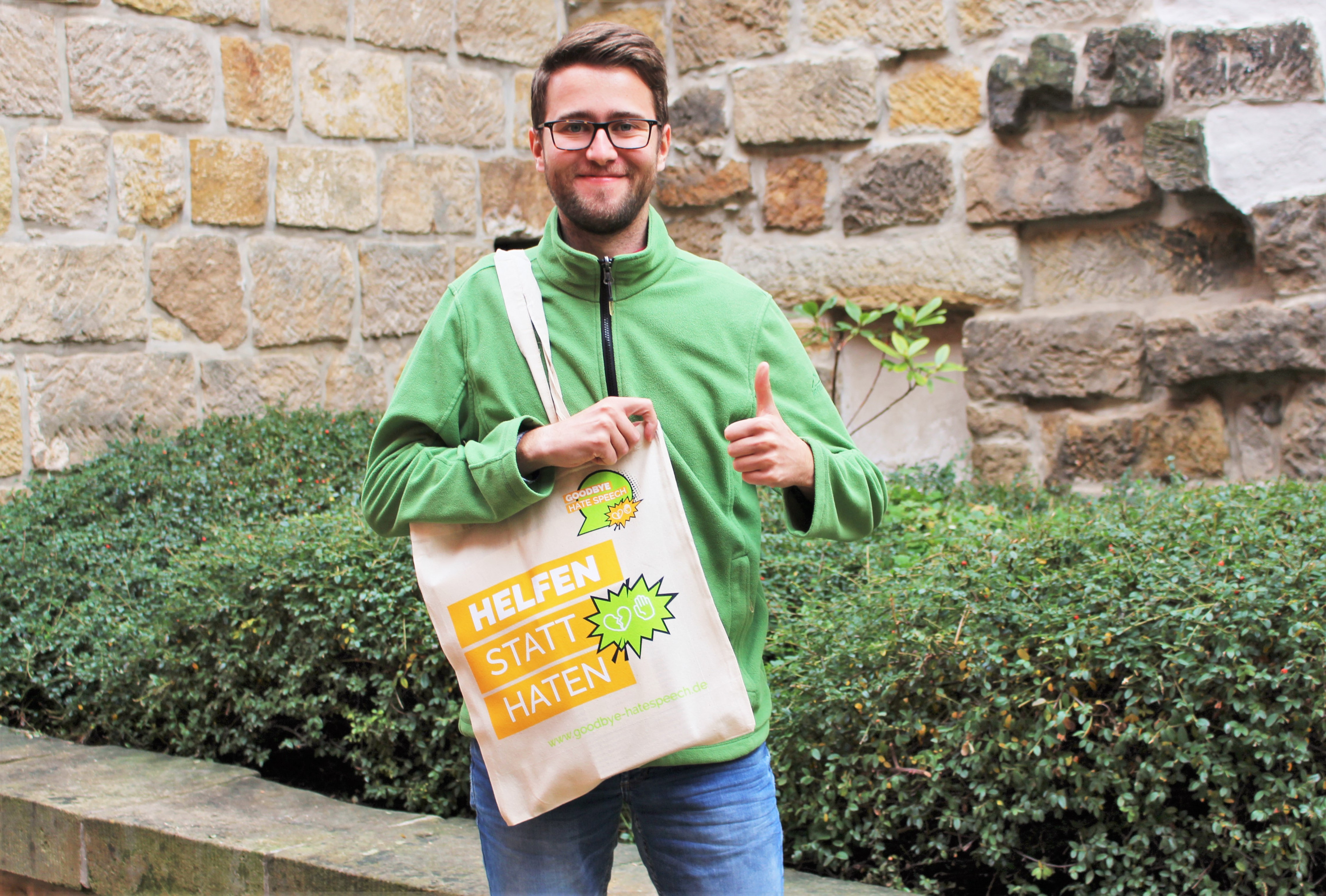 KI generiert: Das Bild zeigt einen lächelnden Mann mit Brille, der einen grünen Pullover trägt und einen Beutel mit der Aufschrift "HELFEN STATT HATEN" in der Hand hält, während er ein Daumen-hoch-Zeichen gibt. Der Hintergrund besteht aus einer Steinmauer und grünen Büschen.