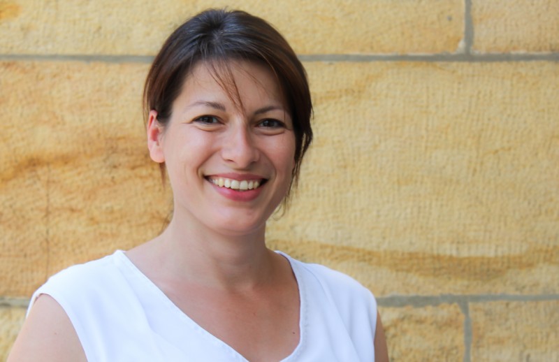 KI generiert: Das Bild zeigt eine lächelnde Frau mit kurzen, dunklen Haaren, die vor einer hellbraunen, steinernen Wand steht. Sie trägt ein weißes Oberteil.