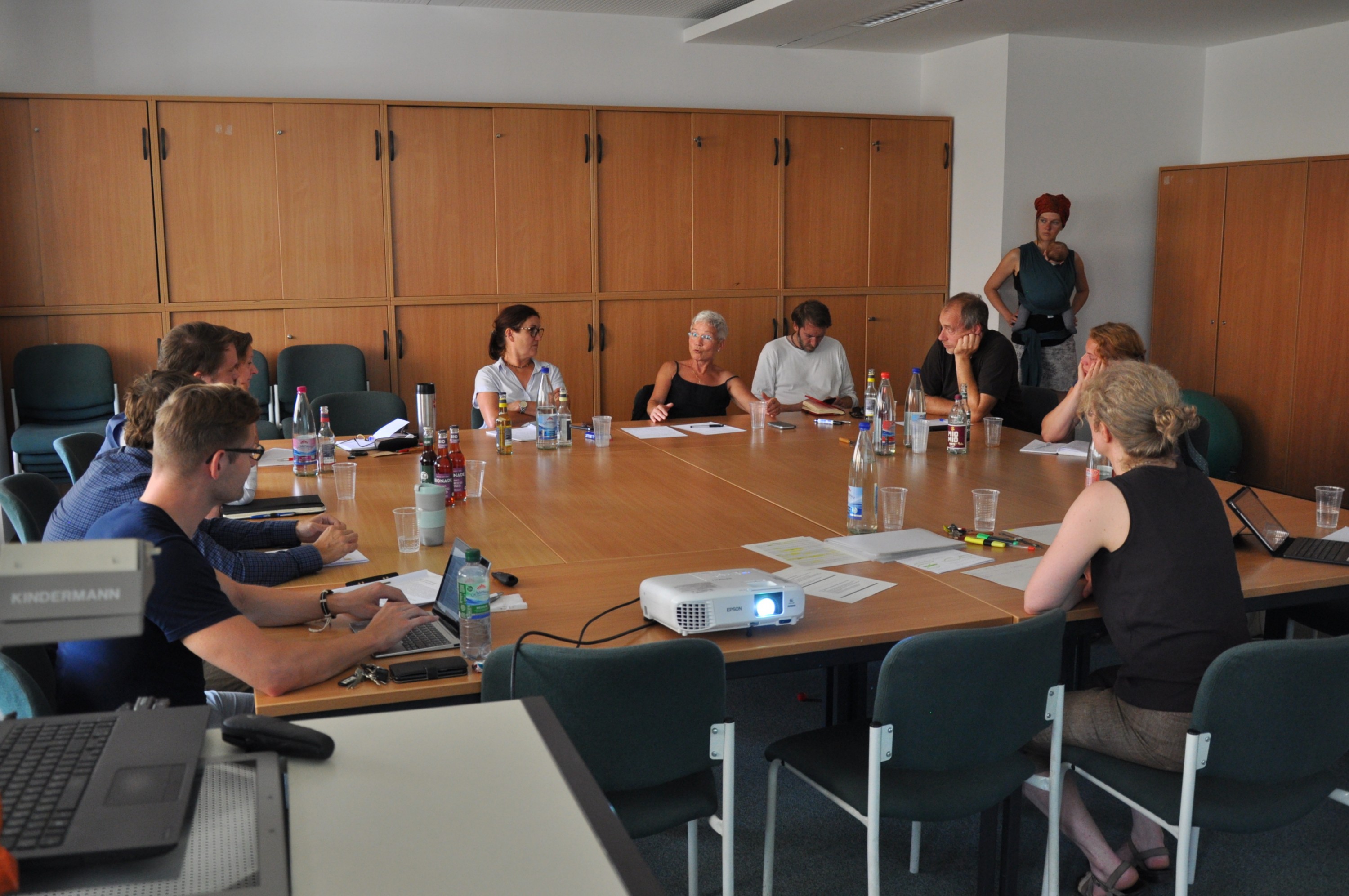 KI generiert: Das Bild zeigt eine Gruppe von Personen, die in einem Besprechungsraum an einem großen ovalen Tisch sitzen und sich in einer Besprechung oder einem Meeting befinden. Der Hauptinhalt des Bildes ist eine formelle Zusammenkunft oder Diskussion in einem Büroumfeld.