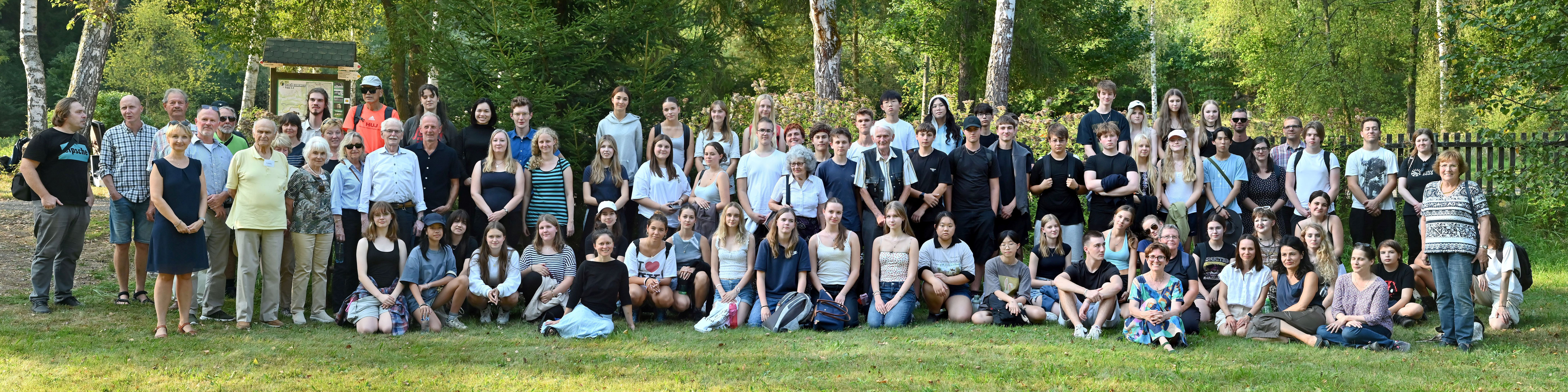 KI generiert: Das Bild zeigt eine große Gruppe von Menschen, die in einem parkähnlichen Umfeld posieren. Der Hauptinhalt ist eine Gruppenfotografie mit Personen verschiedenen Alters, die gemeinsam ein Foto machen.