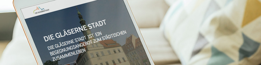 KI generiert: Das Bild zeigt ein Tablet, das eine Webseite mit dem Titel "DIE GLÄSERNE STADT" anzeigt, was auf ein Projekt oder eine Initiative hinweist, die sich mit dem urbanen Leben und der Begegnung in der Stadt befasst. Im Hintergrund sind ein gemütliches Wohnzimmer mit einem Sofa und einer Decke zu sehen.