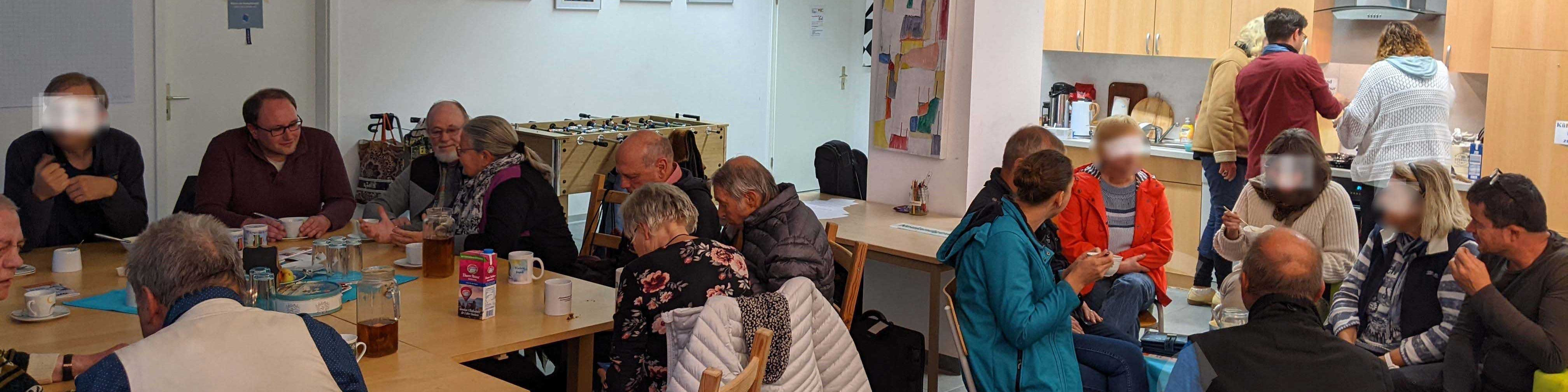 KI generiert: Das Bild zeigt eine Gruppe von Menschen, die in einem Gemeinschaftsraum zusammen sitzen, essen und sich unterhalten. Im Hintergrund sind einige Personen in einer Küche zu sehen, die sich ebenfalls unterhalten und möglicherweise Essen vorbereiten.