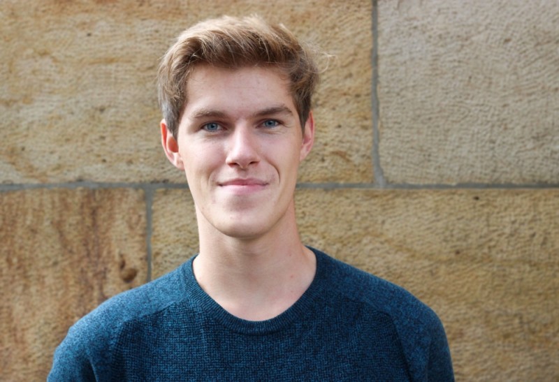 KI generiert: Das Bild zeigt einen jungen Mann mit hellem Haar und blauen Augen, der ein dunkles, langärmliges Oberteil trägt und vor einer steinernen Wand steht. Der Hauptinhalt des Bildes ist das Porträt des lächelnden Mannes.