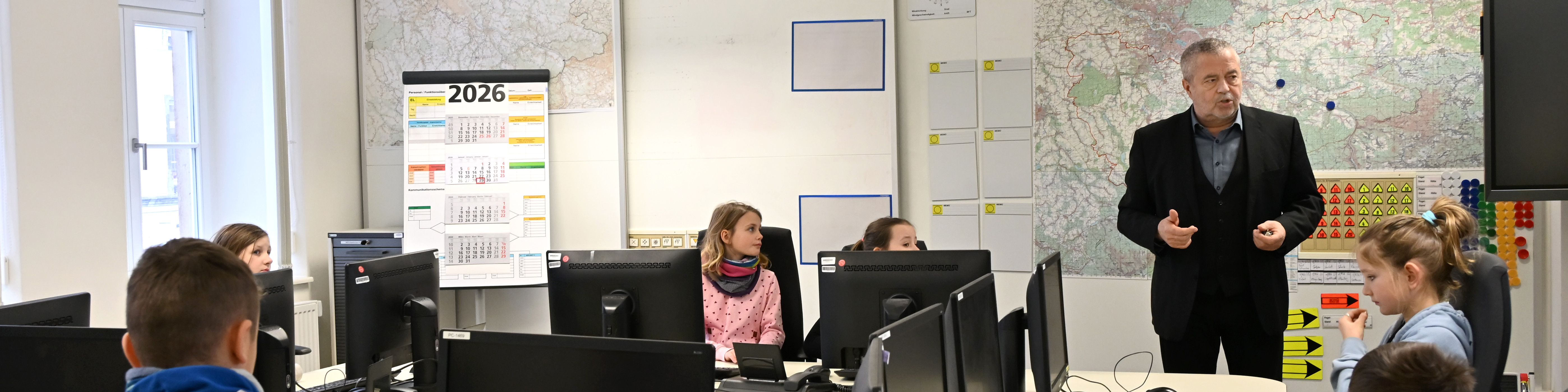 KI generiert: Das Bild zeigt einen Klassenraum, in dem ein Lehrer vor einer Gruppe von Schülern steht und unterrichtet. Die Schüler sitzen an Computern und scheinen einer Präsentation oder einem Vortrag zu folgen.