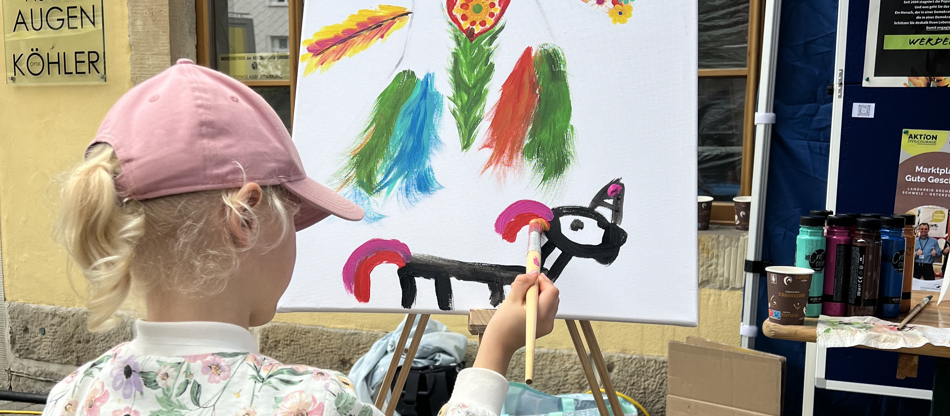 KI generiert: Das Bild zeigt ein Kind mit einer pinken Mütze, das an einem Staffelei steht und ein buntes Bild mit Blumen und einem schwarzen Tier malt. Der Hauptinhalt des Bildes ist das Kind, das kreativ tätig ist und ein Kunstwerk erstellt.