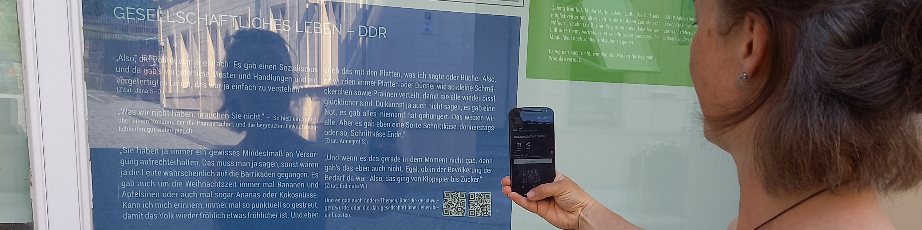 KI generiert: Das Bild zeigt eine Person, die mit einem Smartphone einen QR-Code scannt, der auf einem Plakat an einer Glaswand zu sehen ist. Der Hauptinhalt des Bildes ist das Scannen des QR-Codes, der wahrscheinlich zu weiteren Informationen über das Thema des Plakats führt.