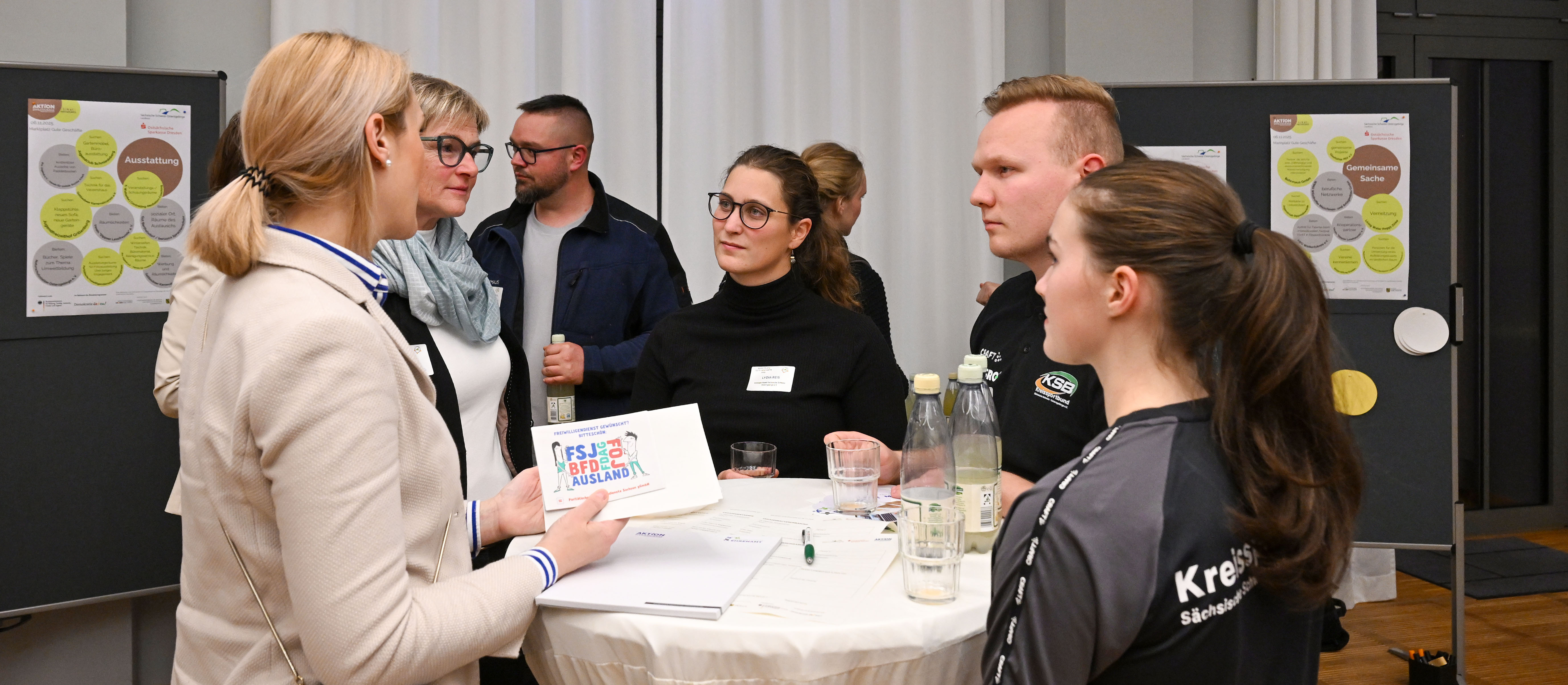 KI generiert: Das Bild zeigt eine Gruppe von Personen, die sich um einen Tisch versammelt haben und sich in einer Diskussion oder einem Meeting befinden. Der Hauptinhalt des Bildes ist eine interaktive Gruppenaktivität, möglicherweise eine Arbeitsbesprechung oder ein Workshop.