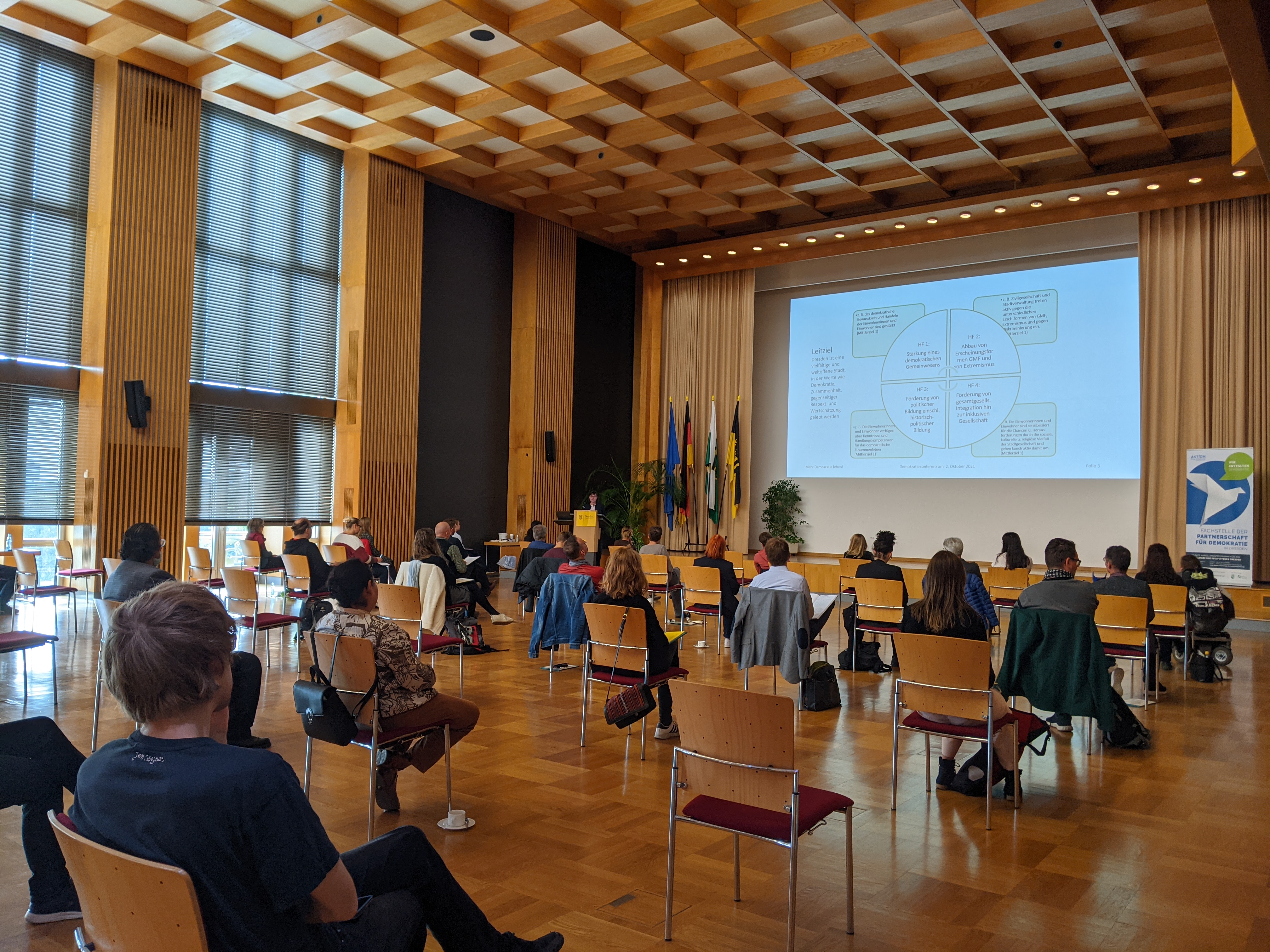 KI generiert: Das Bild zeigt einen Vortragsraum mit mehreren Personen, die auf Stühlen sitzen und einer Präsentation auf einer großen Leinwand folgen. Der Hauptinhalt des Bildes ist eine Konferenz oder ein Seminar in einem modernen, holzvertäfelten Raum mit großen Fenstern.