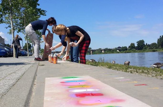 KI generiert: Das Bild zeigt zwei Personen, die mit Sprühdosen bunte Graffiti auf einen Gehweg in der Nähe eines Gewässers sprühen. Im Hintergrund sind weitere Personen, Bäume und ein parkähnliches Umfeld zu sehen.