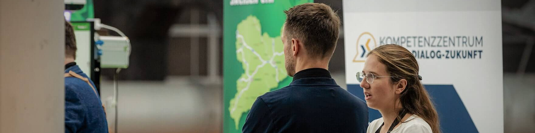 KI generiert: Das Bild zeigt zwei Personen, die sich auf einer Veranstaltung unterhalten, während im Hintergrund ein Stand mit einer Karte und dem Logo des "Kompetenzzentrum Dialog-Zukunft" zu sehen ist. Der Hauptinhalt des Bildes ist die Interaktion der Personen in einem professionellen oder informativen Umfeld.