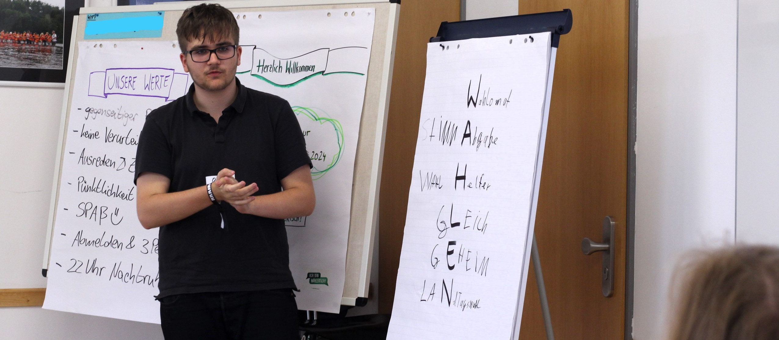 KI generiert: Das Bild zeigt einen Mann, der vor einem Whiteboard und einem Flipchart steht und eine Präsentation hält. Der Hauptinhalt des Bildes ist die Präsentation, bei der Themen wie Werte und Willkommenskultur behandelt werden.