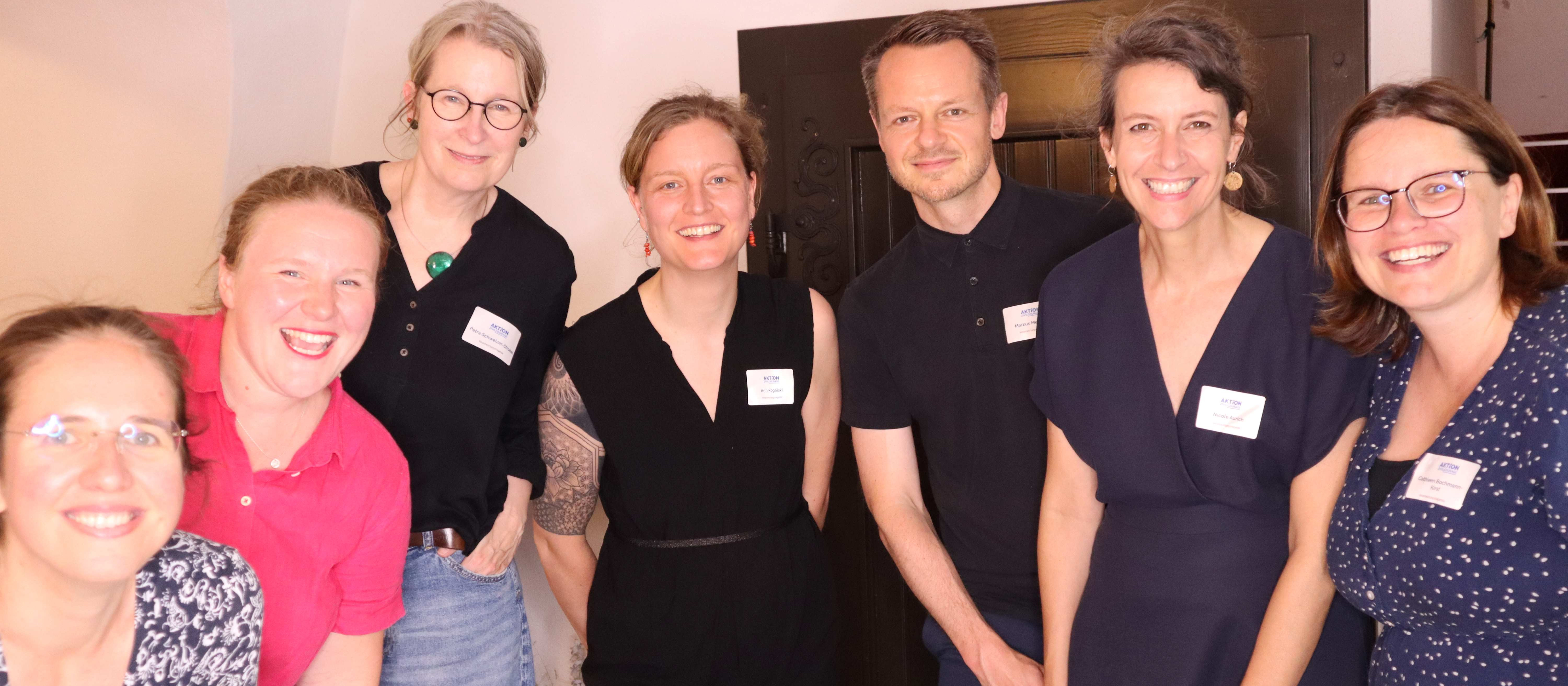 KI generiert: Das Bild zeigt eine Gruppe von sieben Personen, die in einem Innenraum stehen und lächeln. Sie tragen Namensschilder, was darauf hindeutet, dass es sich um eine formelle oder berufliche Veranstaltung handelt.