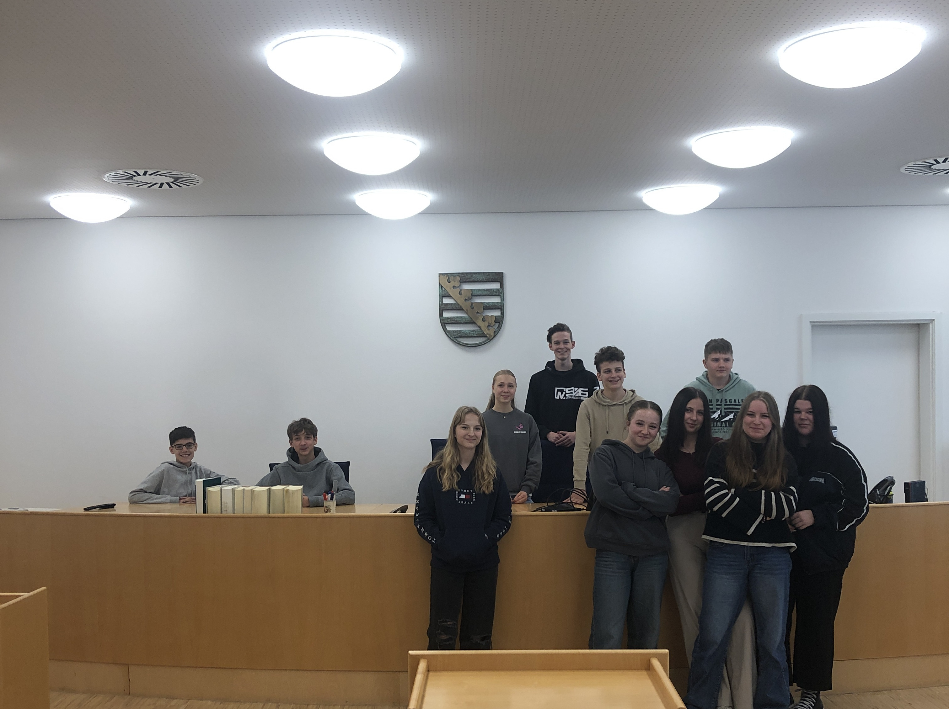 KI generiert: Das Bild zeigt eine Gruppe von Jugendlichen in einem Gerichtssaal, die sich vor einem Richtertisch mit Holzvertäfelung und einem Wappen an der Wand versammelt haben. Der Hauptinhalt des Bildes ist die Darstellung einer Schulklasse oder Jugendgruppe, die möglicherweise an einer Führung oder einem Bildungsprogramm im Gericht teilnimmt.