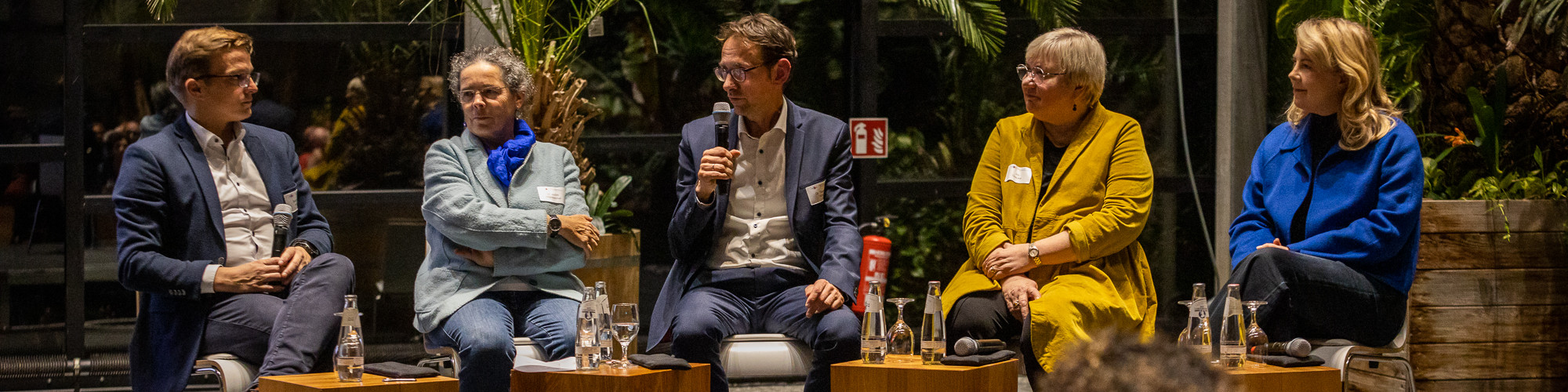 KI generiert: Das Bild zeigt fünf Personen, die auf einer Bühne sitzen und an einer Diskussion oder einem Panel teilnehmen. Der Hauptinhalt des Bildes ist eine öffentliche Veranstaltung oder ein Forum, bei dem die Teilnehmer vor einem Publikum sprechen und diskutieren.