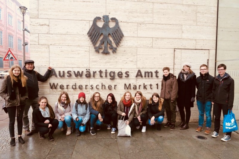 KI generiert: Das Bild zeigt eine Gruppe von Jugendlichen und einem Erwachsenen vor dem Gebäude des Auswärtigen Amts in Berlin. Sie posieren gemeinsam für ein Foto vor dem Eingang, über dem das Emblem der Bundesrepublik Deutschland und die Aufschrift "Auswärtiges Amt Werderischer Markt 1" zu sehen sind.