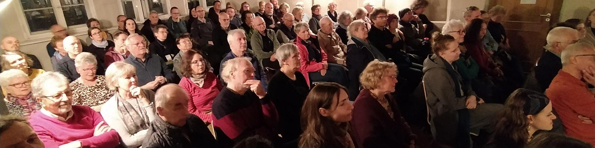KI generiert: Das Bild zeigt eine große Gruppe von Menschen, die in einem Raum sitzen und einer Veranstaltung oder Präsentation zuhören. Der Hauptinhalt des Bildes ist eine aufmerksame Zuhörerschaft in einer Versammlungs- oder Vortragssituation.