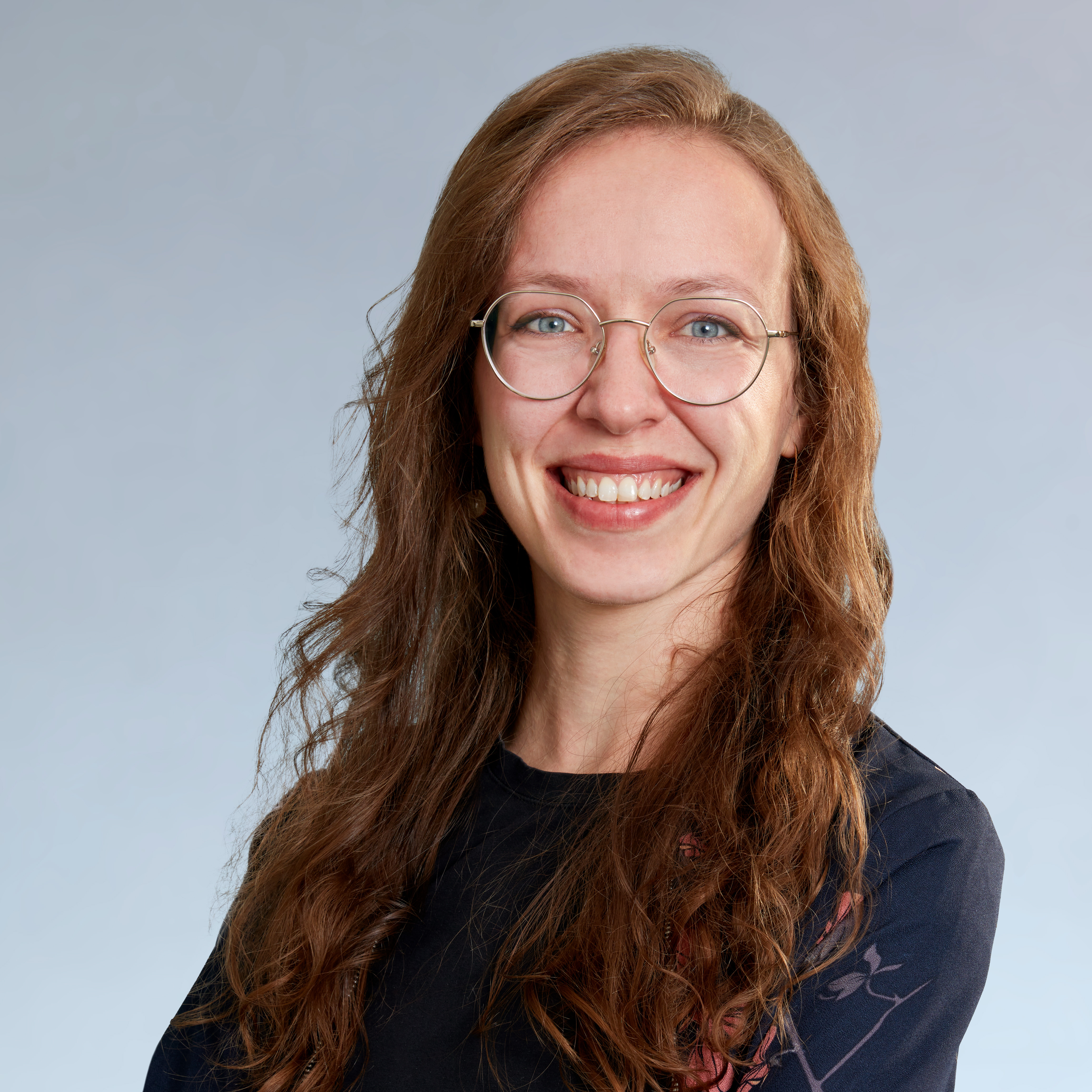 KI generiert: Das Bild zeigt eine lächelnde Frau mit langen, welligen, rötlichen Haaren und einer Brille. Sie trägt ein dunkles Oberteil mit einem Blumenmuster auf der Schulter.