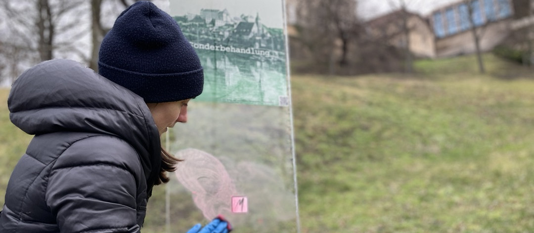 KI generiert: Das Bild zeigt eine Person in Winterkleidung, die eine Informationsafel in einem Freiluftbereich betrachtet. Im Hintergrund sind ein grasbewachsener Hügel und einige Gebäude zu sehen.