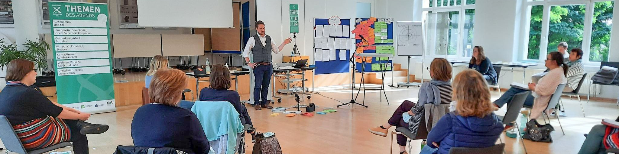 KI generiert: Das Bild zeigt eine Gruppe von Personen in einem hellen Raum, die einem Vortragenden zuhören, der vor einem Flipchart mit bunten Notizen steht. Der Hauptinhalt des Bildes ist eine Präsentation oder ein Workshop in einer informellen Lernumgebung.