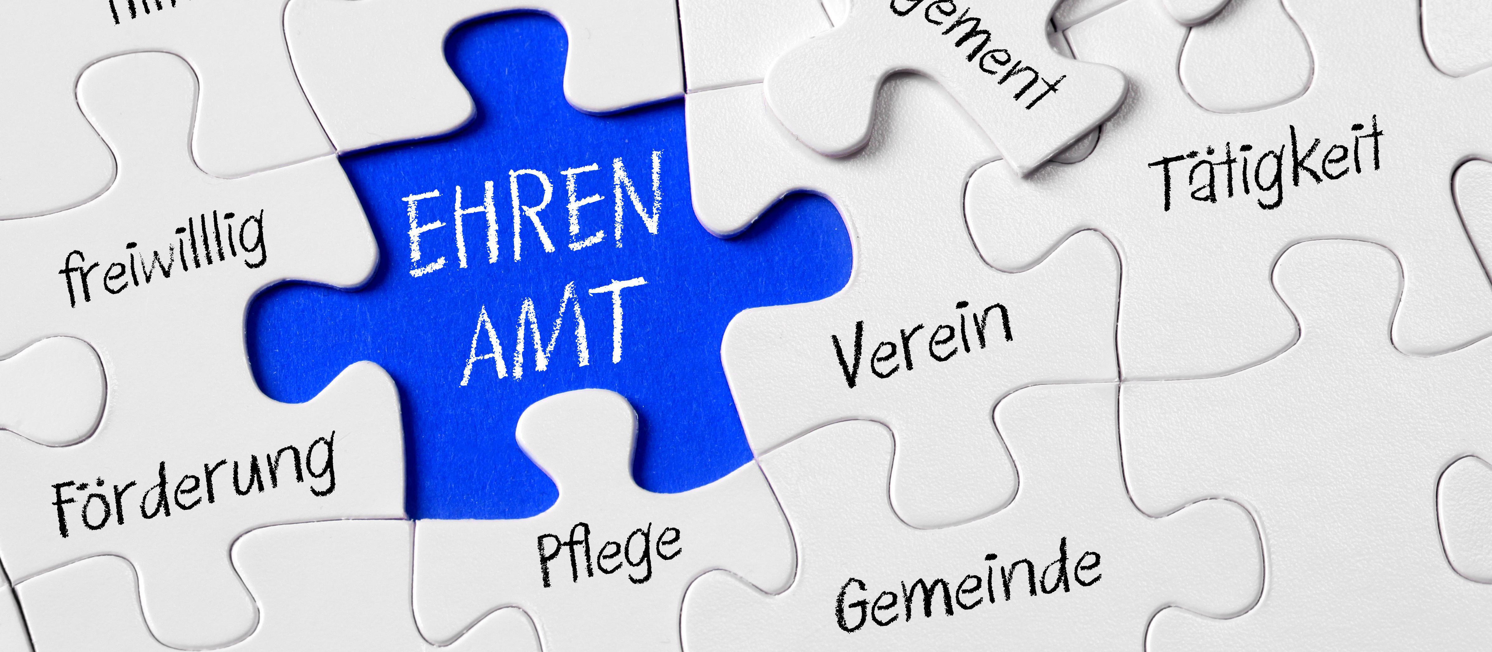 KI generiert: Das Bild zeigt ein Puzzle, bei dem ein blaues Puzzleteil mit der Aufschrift "EHREN AMT" im Mittelpunkt steht. Um dieses zentrale Puzzleteil herum sind weitere weiße Puzzleteile mit Begriffen wie "freiwillig", "Förderung", "Pflege", "Gemeinde", "Verein", "Engagement" und "Tätigkeit" angeordnet.