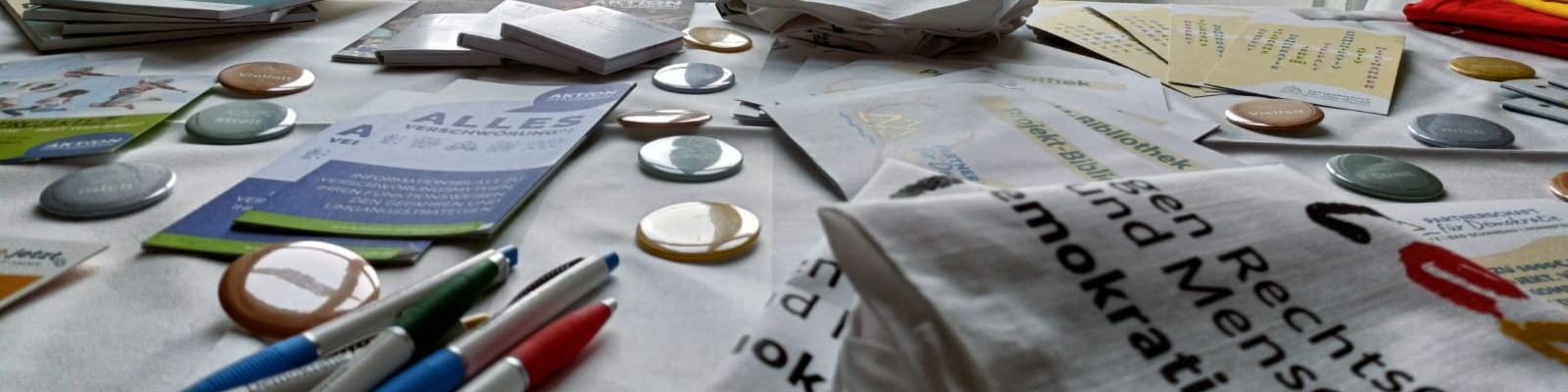 KI generiert: Das Bild zeigt einen Tisch mit verschiedenen Materialien wie Flyern, Buttons, Stiften und einem Beutel, die möglicherweise für eine Informationsveranstaltung oder Kampagne vorbereitet wurden. Der Hauptinhalt des Bildes sind diese Materialien, die auf dem Tisch verstreut liegen.