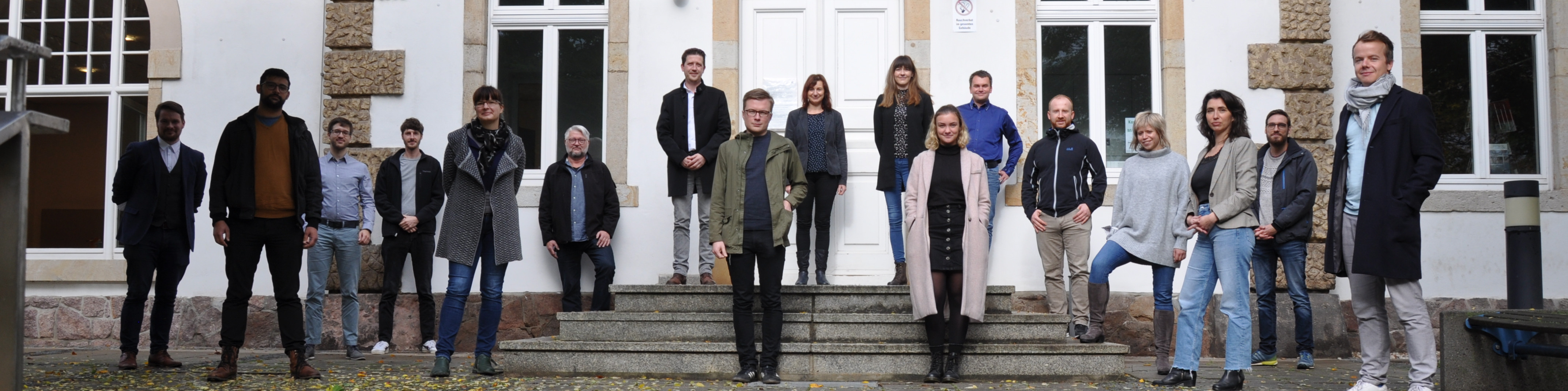 KI generiert: Das Bild zeigt eine Gruppe von 18 Personen, die vor einem Gebäude mit großen Fenstern und einer Treppe posieren. Die Personen sind in verschiedenen Kleidungsstilen gekleidet und scheinen sich für ein Gruppenfoto versammelt zu haben.