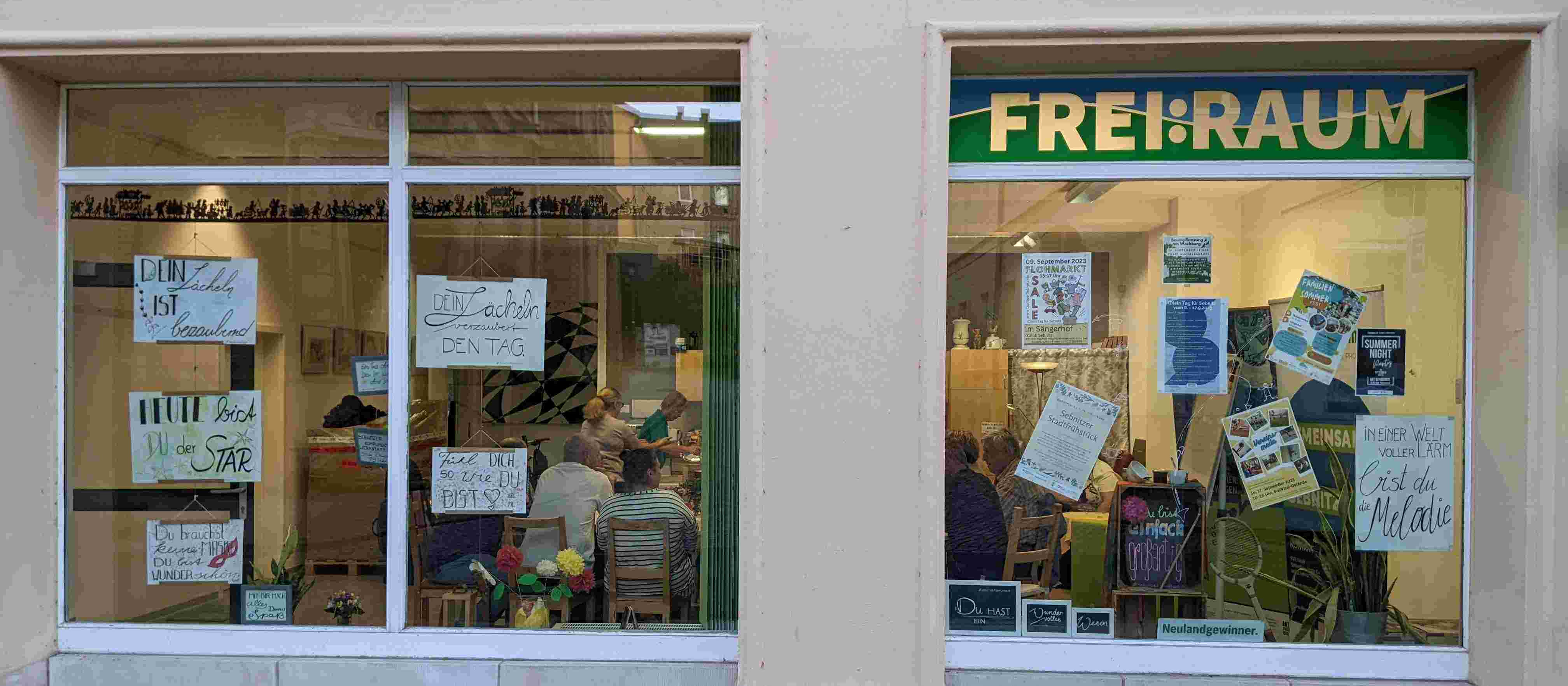 KI generiert: Das Bild zeigt die Schaufenster eines Ladens namens "FREI:RAUM" mit verschiedenen Plakaten und Anzeichen in den Fenstern. Im Inneren sind Menschen zu sehen, die sich unterhalten oder arbeiten.