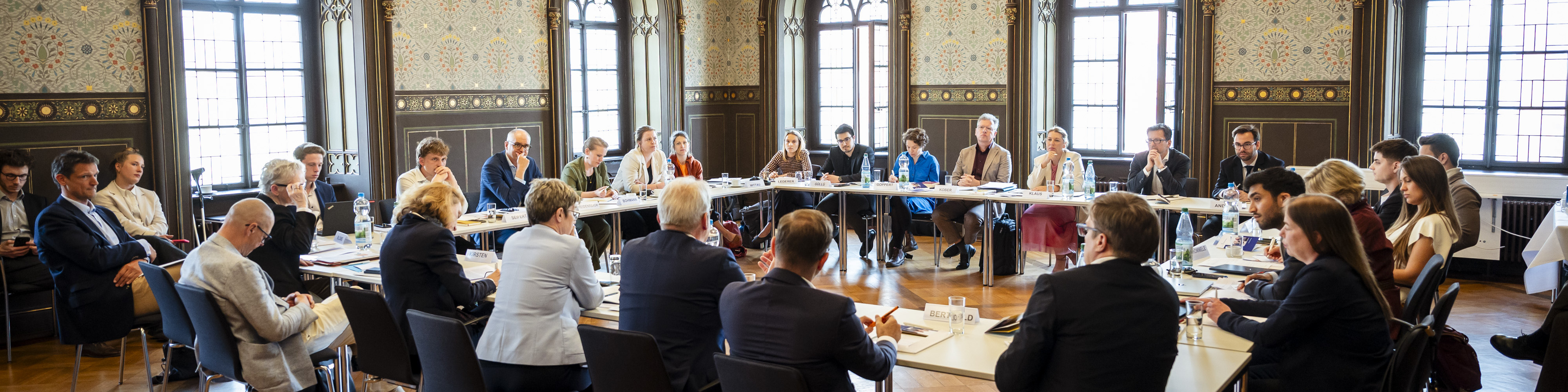 KI generiert: Das Bild zeigt eine formelle Besprechung oder Konferenz in einem historischen Raum mit großen Fenstern und verzierten Wänden. Mehrere Personen sitzen an langen Tischen, die in einem U-förmigen Arrangement angeordnet sind, und scheinen sich auf eine Diskussion oder Präsentation zu konzentrieren.