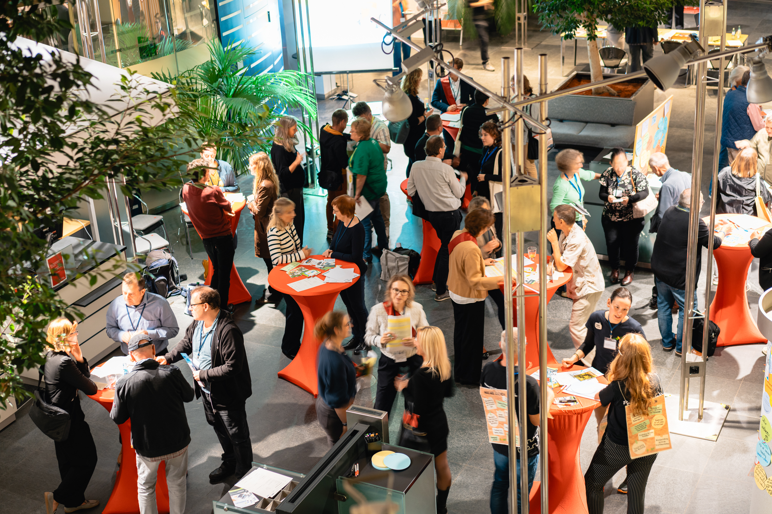 KI generiert: Das Bild zeigt eine belebte Indoor-Veranstaltung mit vielen Menschen, die an hohen Stehtischen stehen und sich unterhalten. Der Hauptinhalt des Bildes ist eine Netzwerk- oder Messeveranstaltung in einer modernen, gut beleuchteten Umgebung mit Pflanzen und verschiedenen Ausstellungsständen.