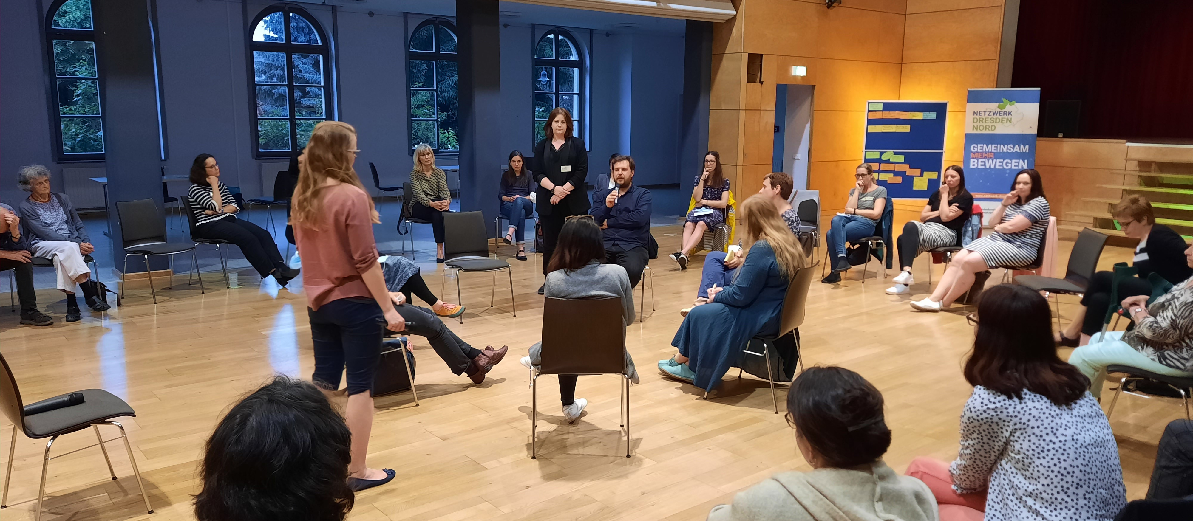 KI generiert: Das Bild zeigt eine Gruppe von Menschen, die in einem Kreis in einem Raum mit Holzboden und großen Fenstern sitzen und sich unterhalten. Es scheint sich um eine Diskussionsrunde oder ein Seminar zu handeln, bei dem die Teilnehmer aktiv teilnehmen und zuhören.