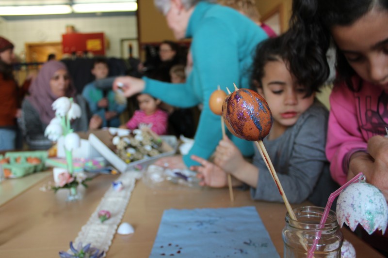 KI generiert: Das Bild zeigt eine Gruppe von Menschen, darunter Kinder und Erwachsene, die gemeinsam an einem Tisch sitzen und Ostereier bemalen. Im Vordergrund sind bunt bemalte Eier auf Spießen und verschiedene Bastelmaterialien zu sehen.