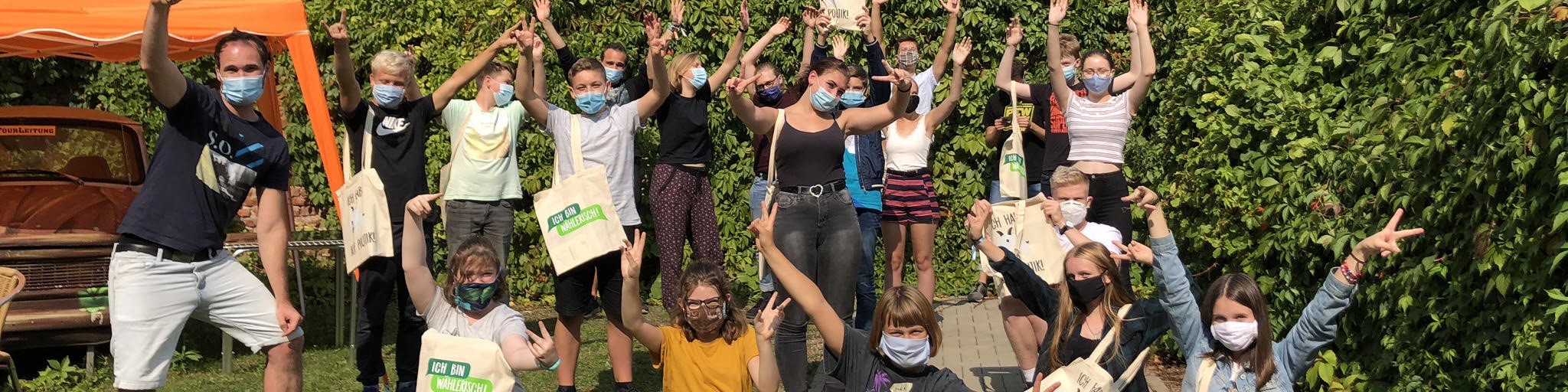 KI generiert: Das Bild zeigt eine Gruppe von Menschen, die draußen vor einer grünen Hecke stehen und sich freudig mit hochgestreckten Armen präsentieren, wobei viele von ihnen Gesichtsmasken tragen. Der Hauptinhalt des Bildes ist eine fröhliche Gruppenaktivität, möglicherweise im Rahmen einer gemeinnützigen oder ökologischen Veranstaltung.
