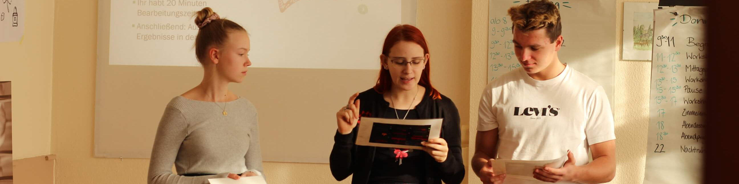 KI generiert: Das Bild zeigt drei Personen, die in einem Raum stehen und jeweils ein Blatt Papier in den Händen halten. Es scheint sich um eine Besprechung oder Präsentation zu handeln, da im Hintergrund eine Projektionsleinwand mit Text zu sehen ist.