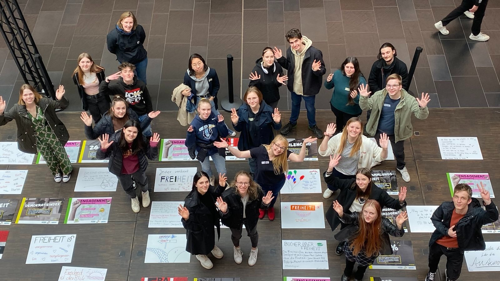 KI generiert: Das Bild zeigt eine Gruppe von Jugendlichen, die fröhlich in die Kamera winken und auf einem Boden mit verschiedenen Plakaten und Aufschriften stehen. Der Hauptinhalt des Bildes ist die Darstellung einer engagierten und lebhaften Gruppe, die möglicherweise an einem Projekt oder einer Veranstaltung teilnimmt.