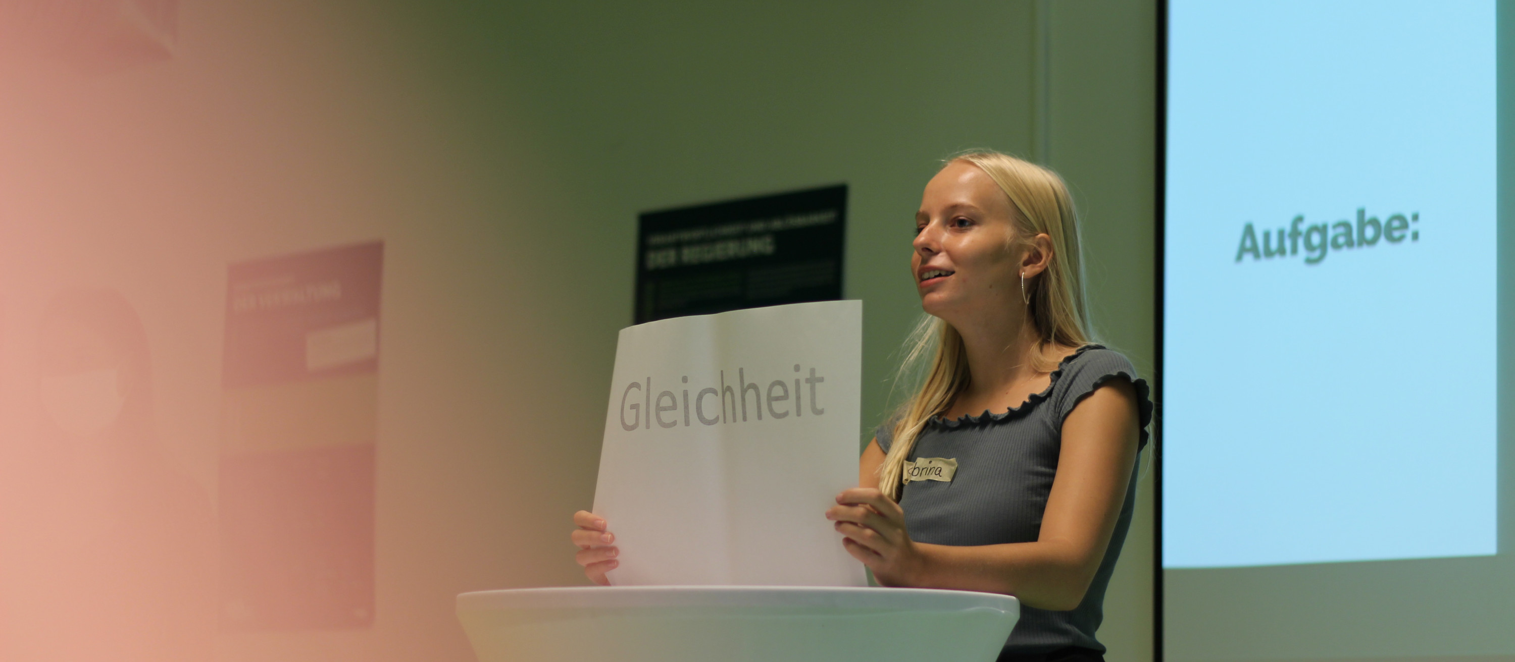 KI generiert: Das Bild zeigt eine Person, die an einem Pult steht und ein Blatt Papier mit der Aufschrift "Gleichheit" hochhält. Im Hintergrund ist eine Leinwand mit dem Wort "Aufgabe:" zu sehen.