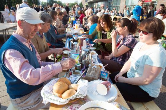 KI generiert: Das Bild zeigt eine belebte Outdoor-Veranstaltung, bei der Menschen an langen Tischen sitzen und gemeinsam essen und trinken. Im Vordergrund bedient ein Mann mit Basecap und Weste die Gäste mit Brot und anderen Speisen.
