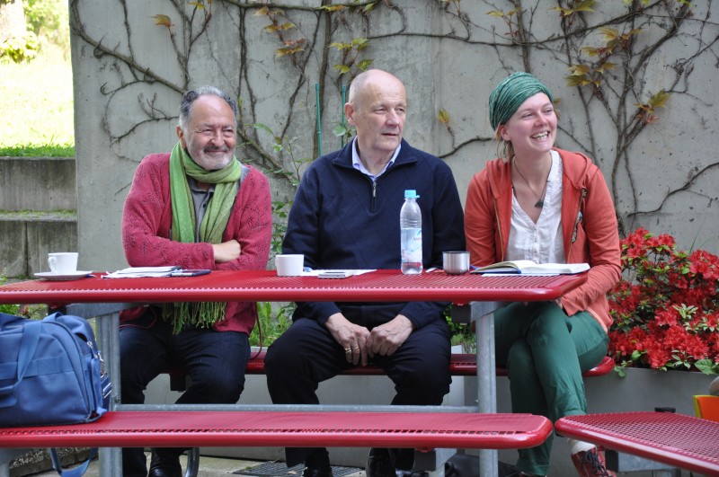KI generiert: Das Bild zeigt drei Personen, die an einem Tisch sitzen und sich unterhalten. Im Vordergrund sind ein Wasserflasche und einige Tassen zu sehen, während im Hintergrund eine Wand mit Kletterpflanzen und Blumen zu erkennen ist.