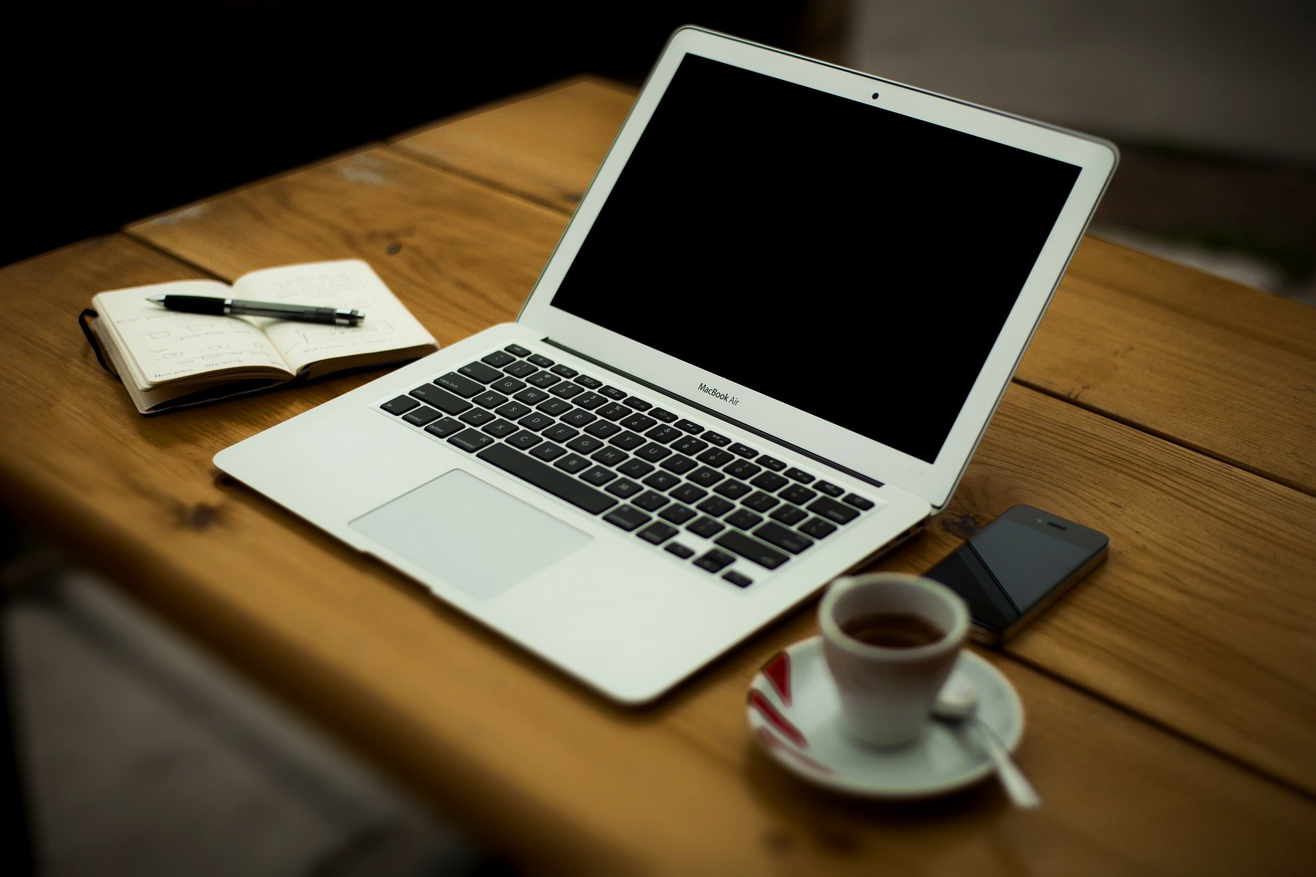 KI generiert: Das Bild zeigt einen Arbeitsplatz mit einem Laptop, einem Notizbuch und einem Stift auf einem Holztisch. Daneben stehen eine Tasse Kaffee und ein Smartphone. Der Hauptinhalt des Bildes ist ein organisierter Arbeitsbereich.