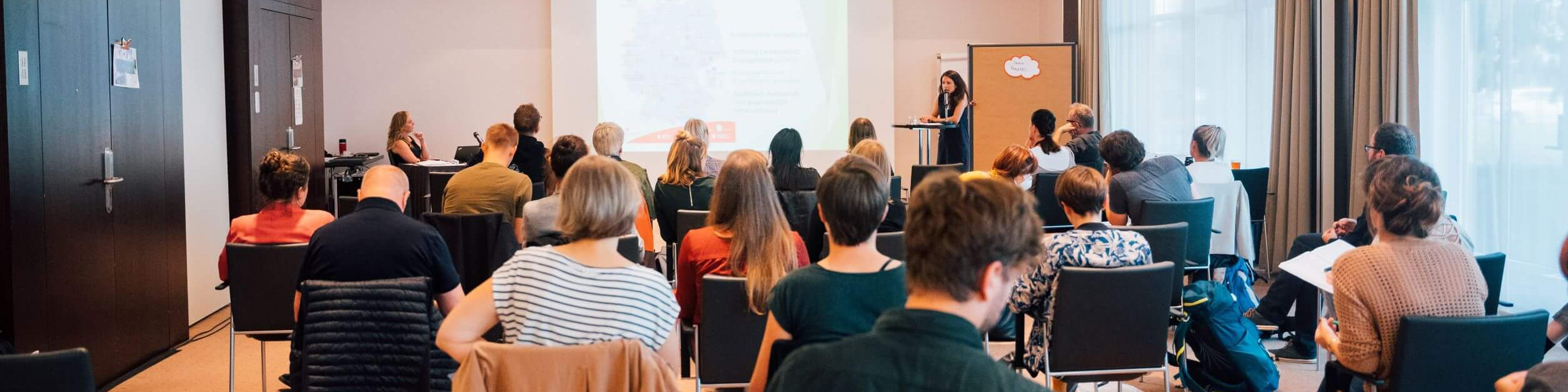 KI generiert: Das Bild zeigt eine Gruppe von Personen in einem Seminarraum, die einer Vortragenden zuhören, die an einem Pult steht. Der Raum ist hell und modern eingerichtet, mit großen Fenstern und einer Projektionsfläche im Hintergrund.